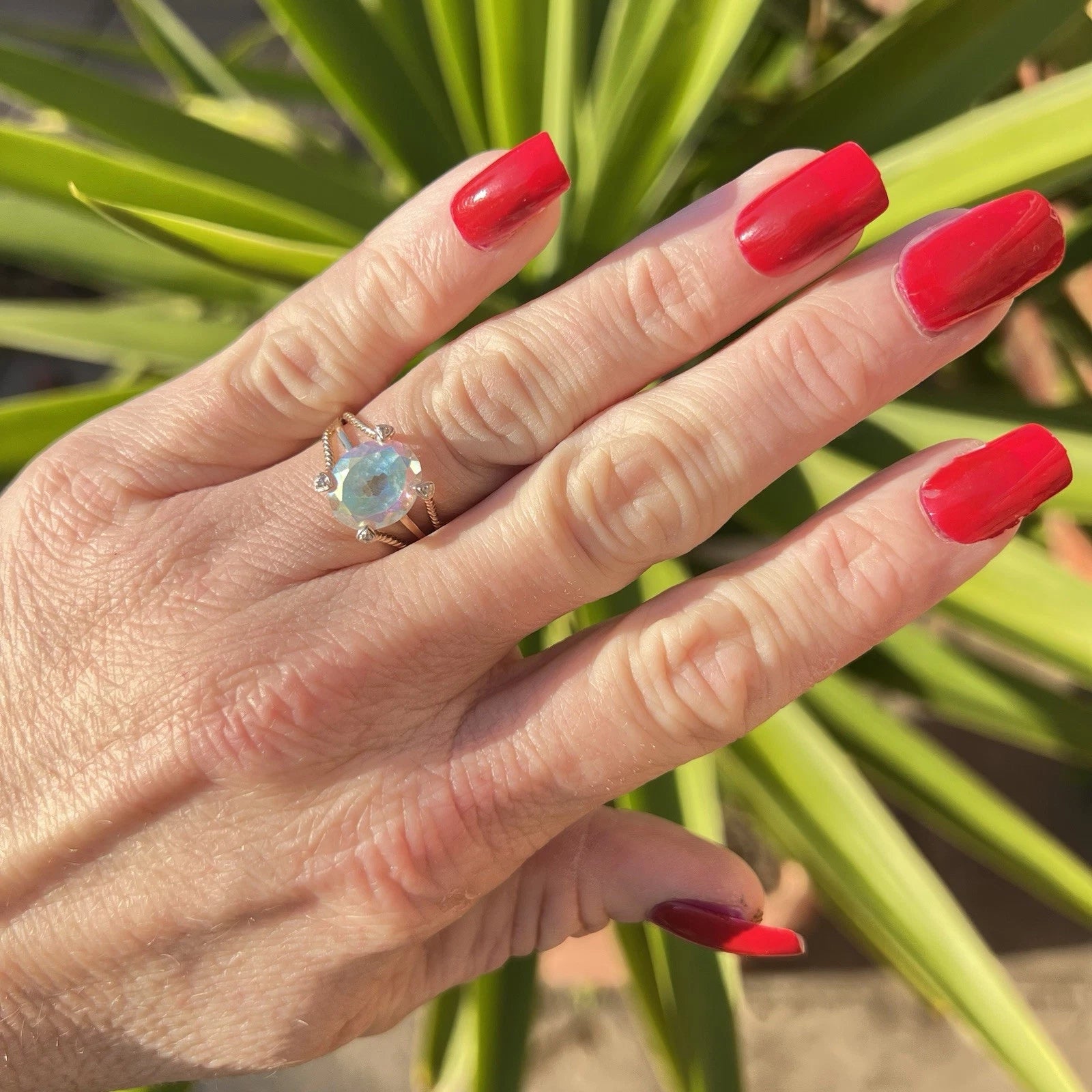 Hand with red nail polish wearing a mystic quartz gold ring against a plant background