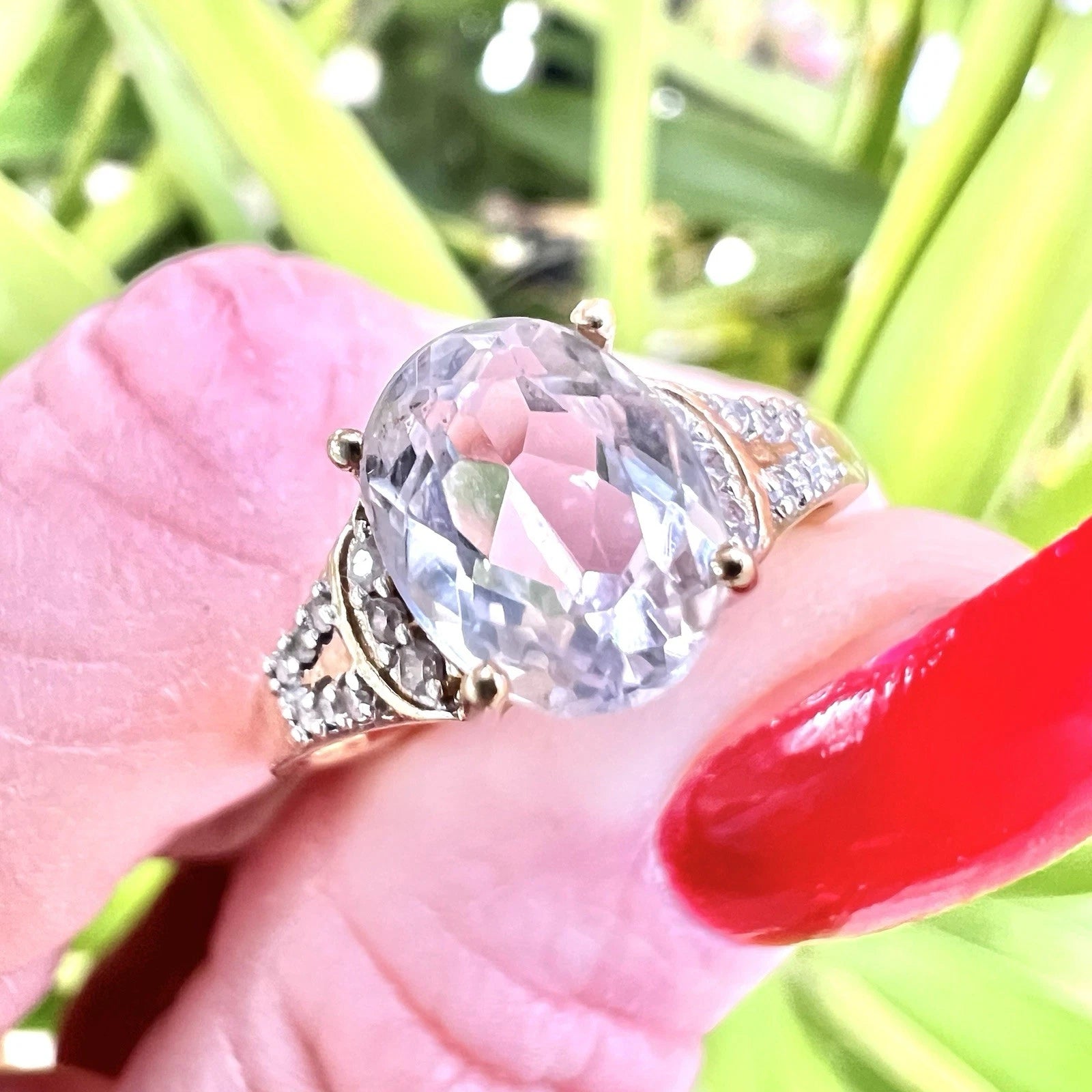 Gold ring with a large clear quartz and diamond gemstones being held by on a hand with red nail polish, green leaves in the background