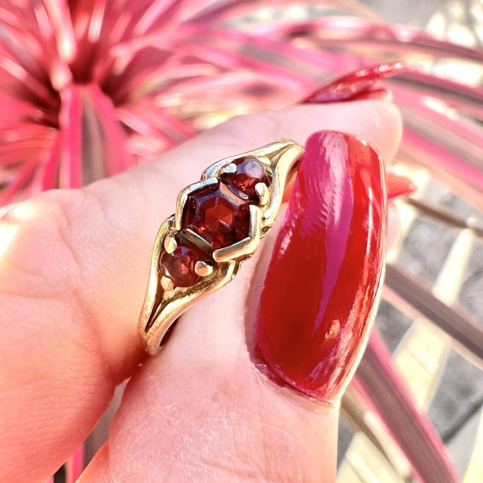 Gold ring with red gemstone on a finger against a blurred pink floral background