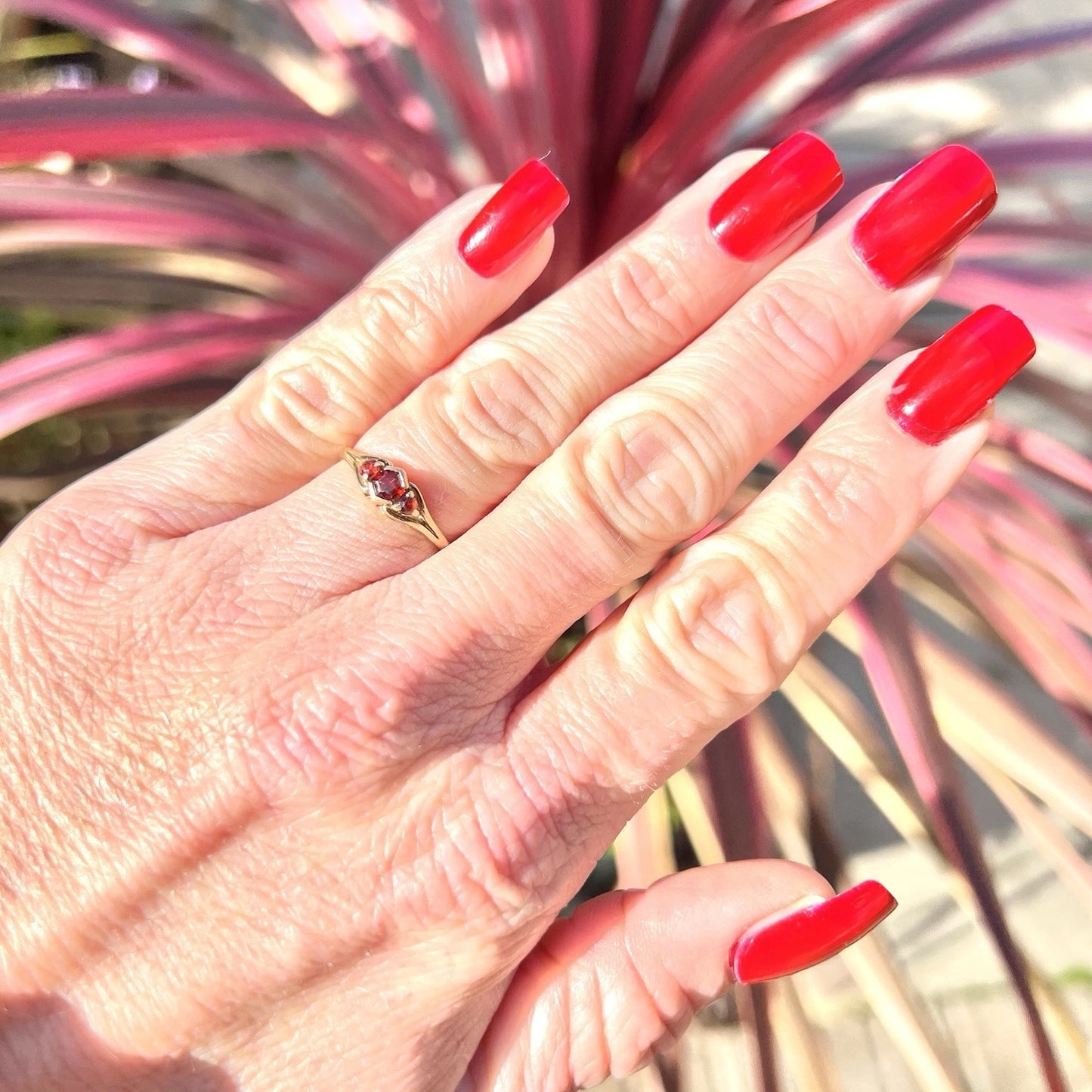 Hand with red nail polish and a ring, against a blurred plant background