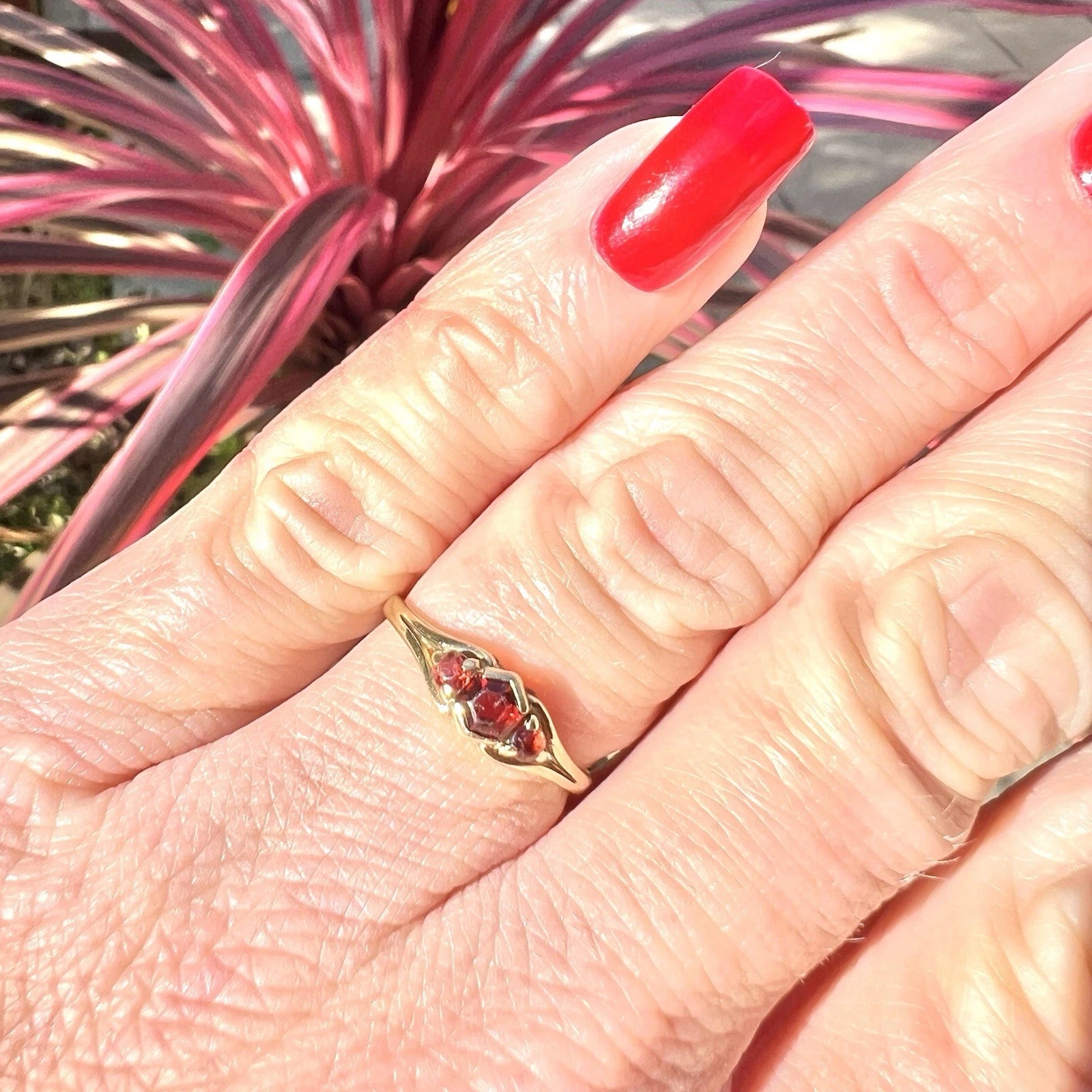 Hand wearing a gold ring with a red gemstone in front of pink and green foliage