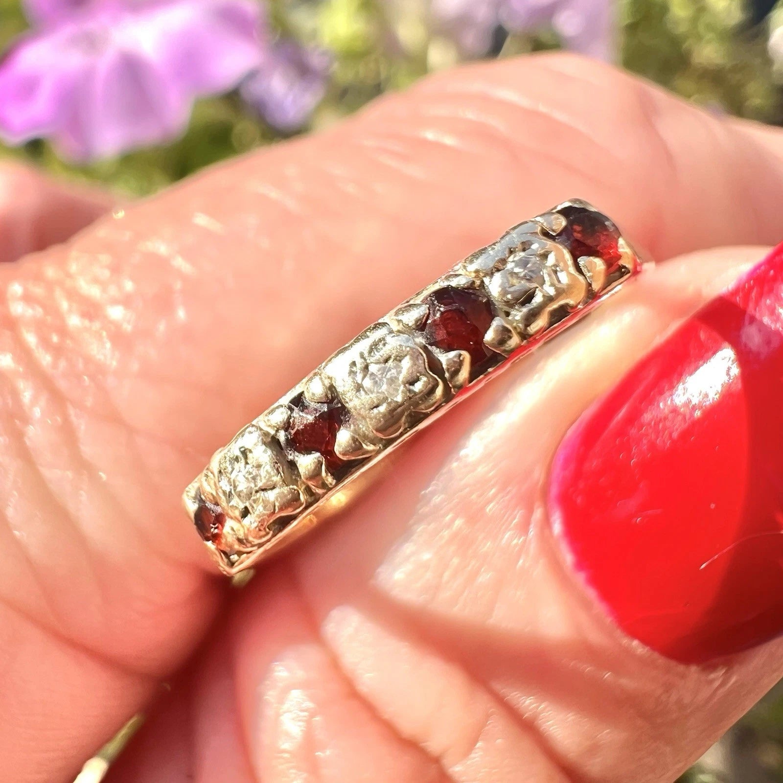 Gold ring with red ruby and cubic zirconia stones being held in fingers with red nail polish against a blurred floral background