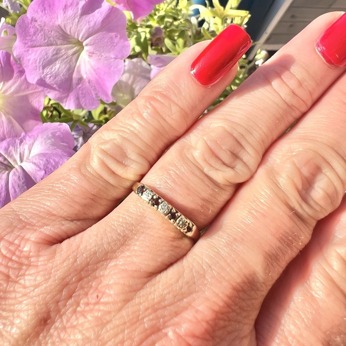 Hand wearing a gold ring with red rubies and cubic zirconia, red nail polish, and purple flowers in the background