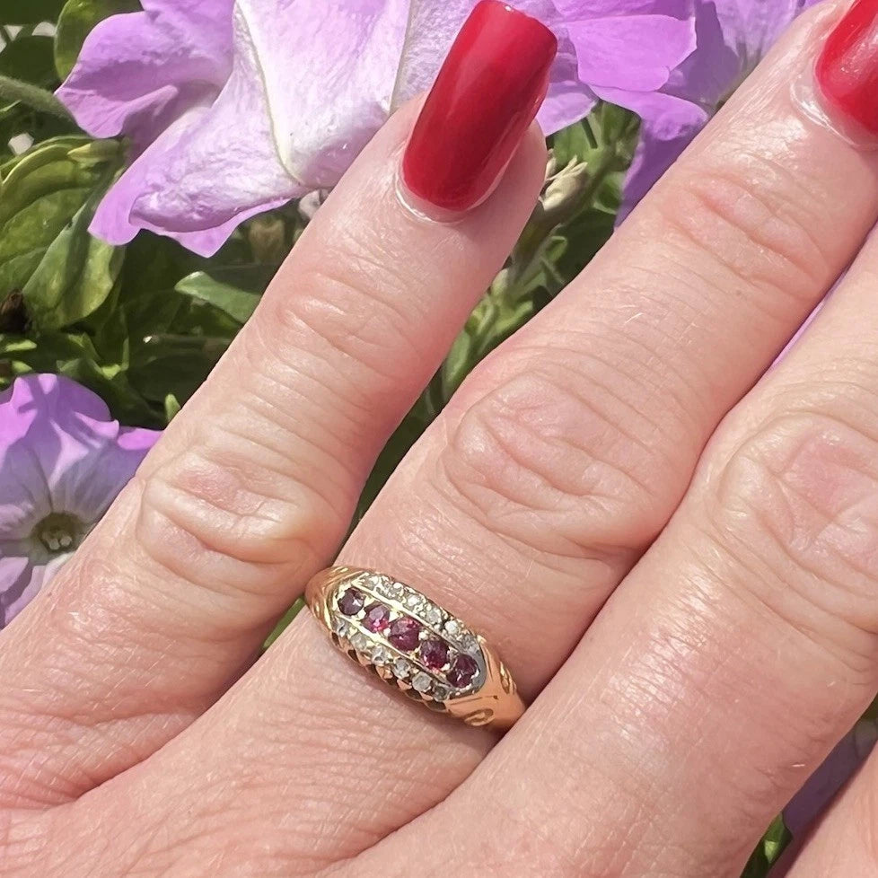 Close-up of a hand wearing a gold ring with red rubies and diamond against a background of purple flowers.