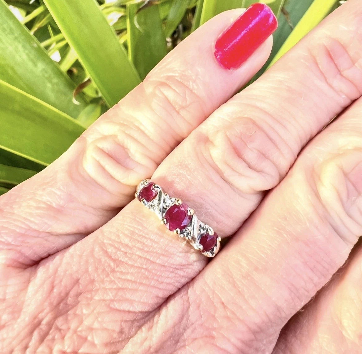 Ring with red ruby gemstones and diamonds on a finger against a green leafy background