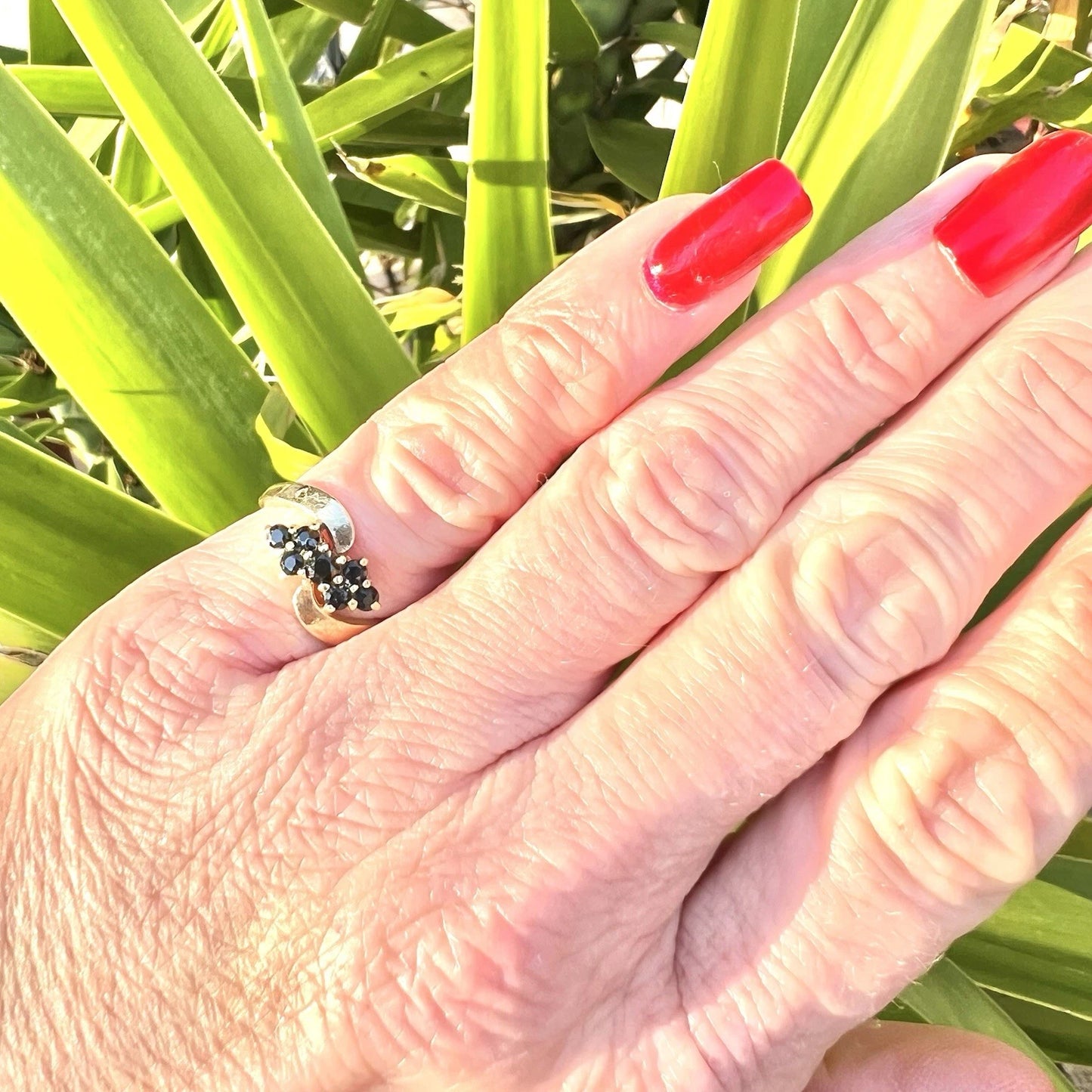Hand with red nail polish wearing a sapphire cluster ring against a green leafy background
