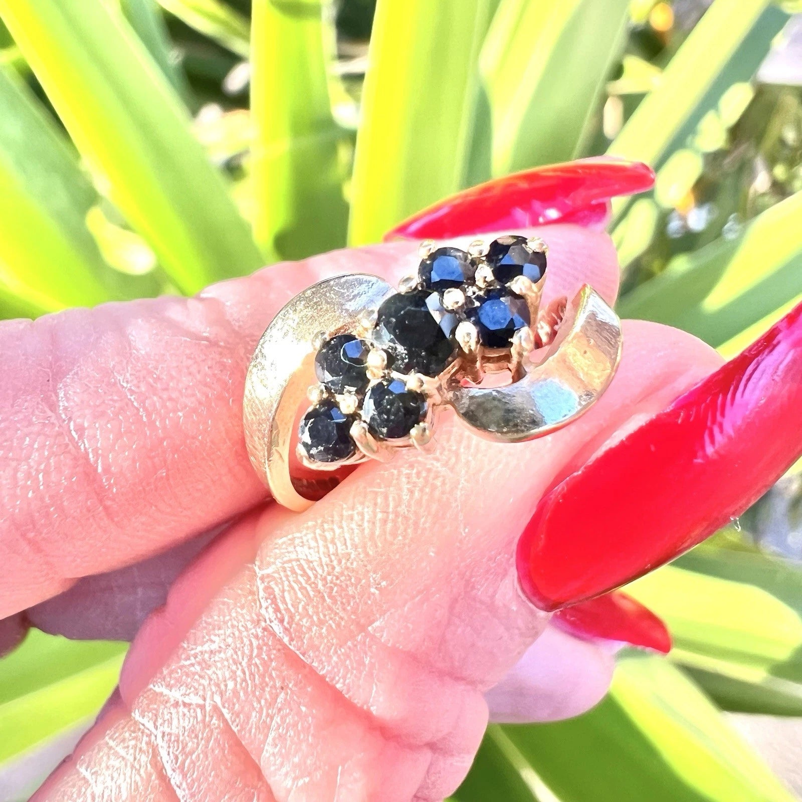 Gold ring with sapphire gemstones held by a hand with red nail polish against a green plant background
