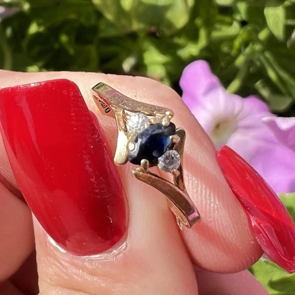 Gold ring with a blue sapphire gemstone on a finger with red nail polish against a blurred natural background