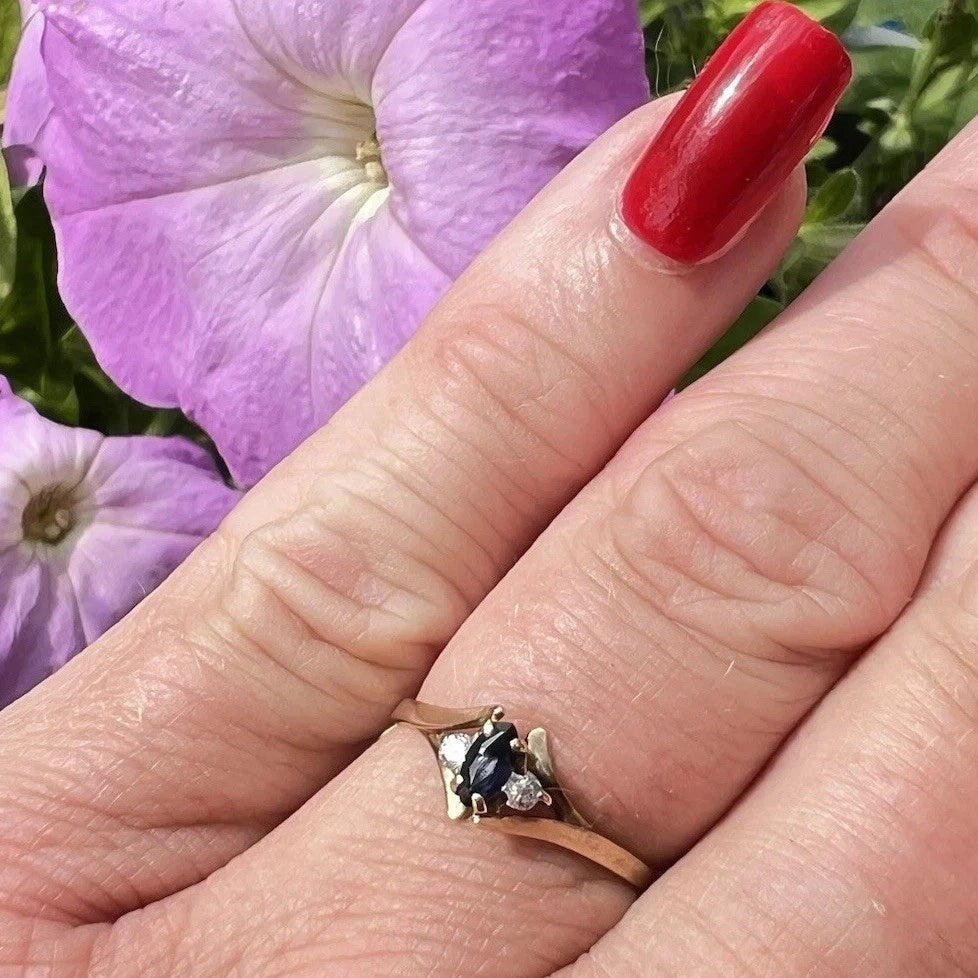 Hand wearing a gold ring with a blue sapphire gemstone against a purple flower background