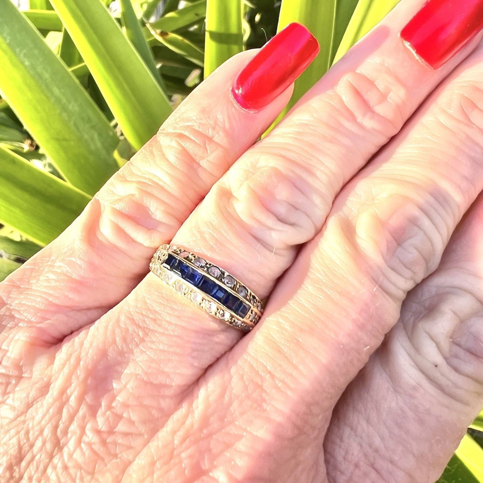 Hand wearing a ring with blue sapphire  gemstones against a green leafy background
