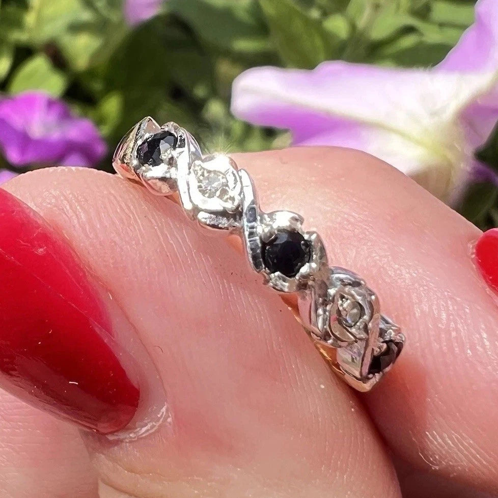 Gold ring with alternating sapphire and diamonds being held by a finger with red nail polish, against a blurred floral background.