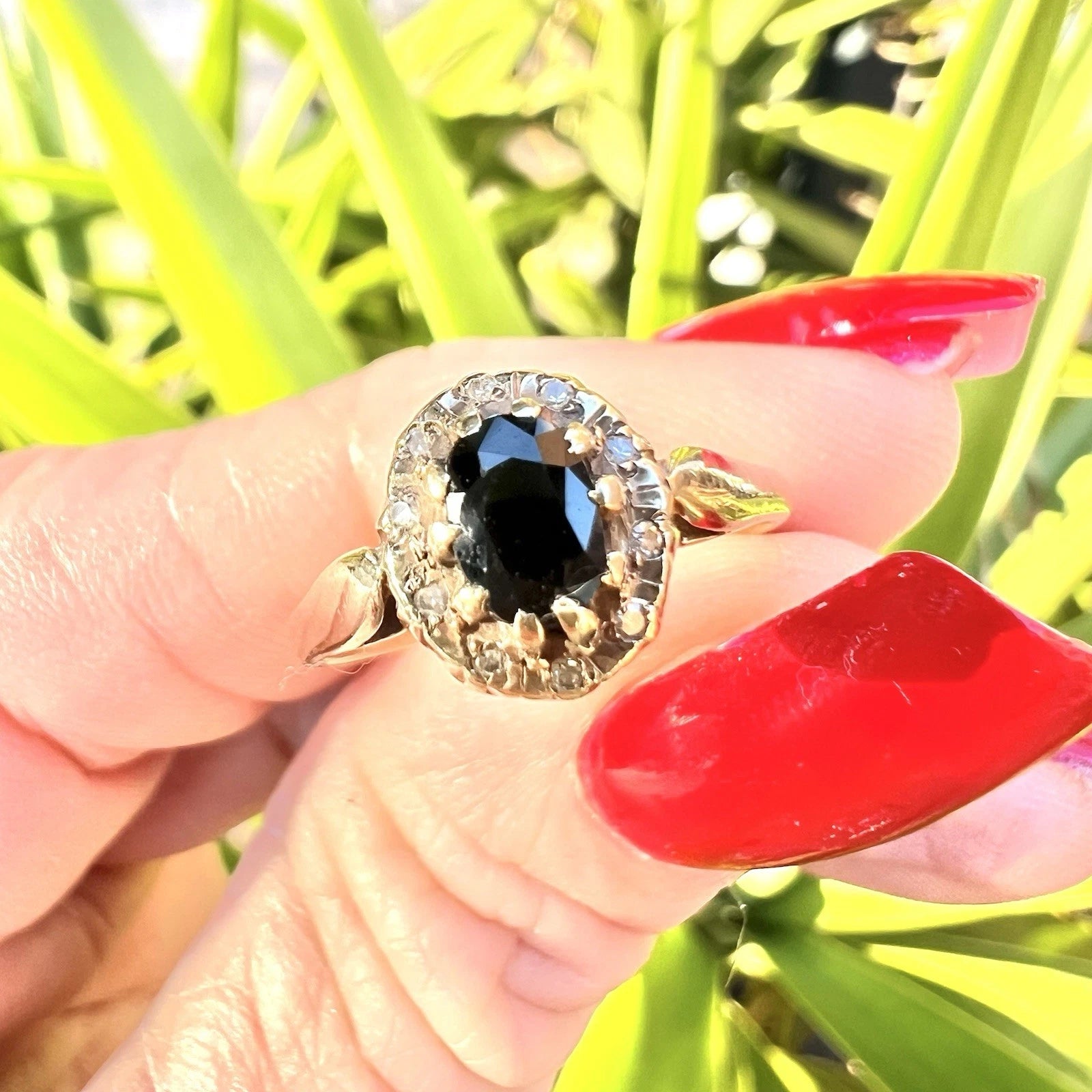 Gold ring with a sapphire gemstone and diamond surround held by a hand with red nail polish against a green background