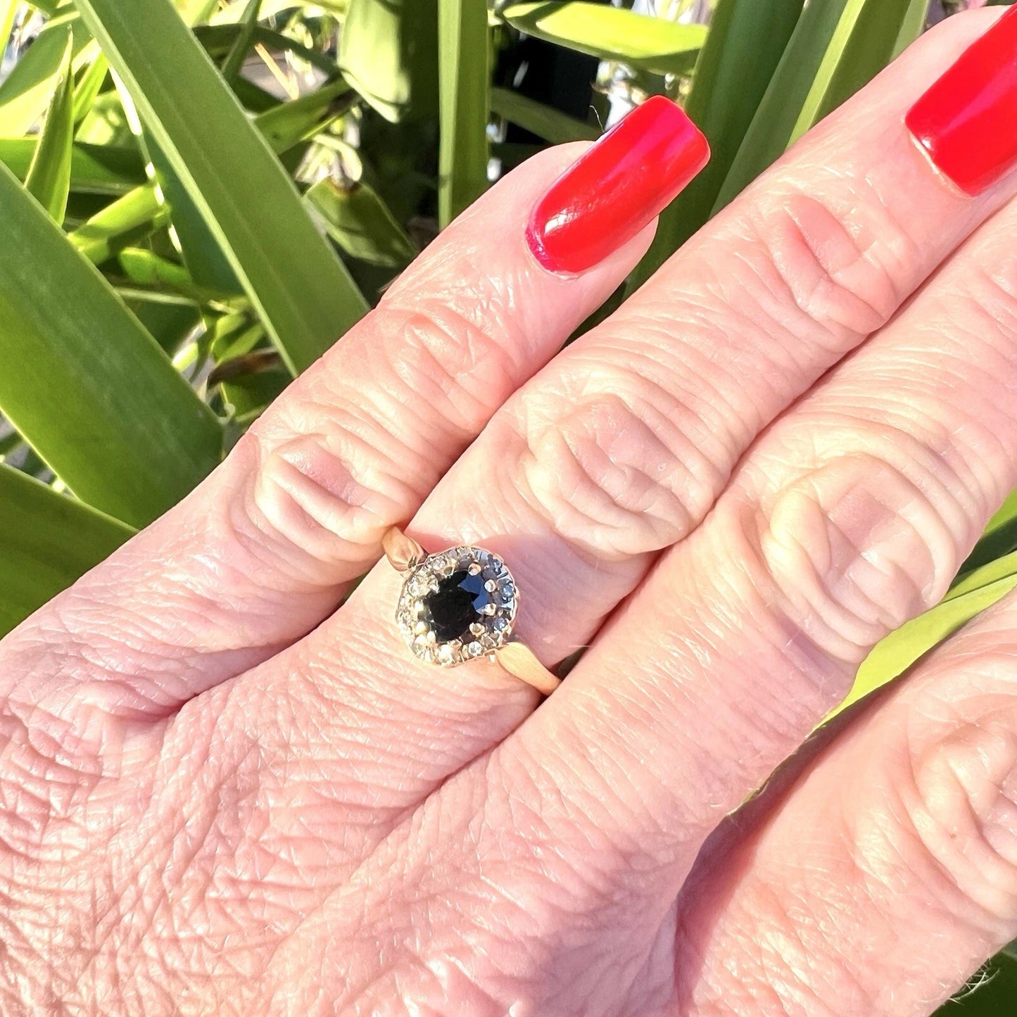 Hand wearing a ring with a blue sapphire and diamond halo against a green leafy background