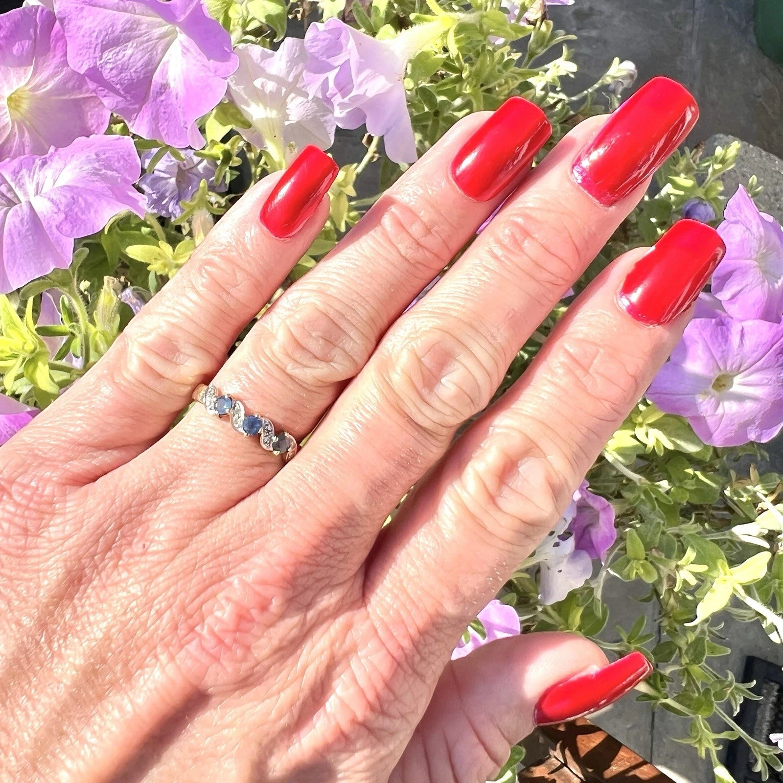 Hand with red nails and a light blue sapphire ring, surrounded by purple flowers