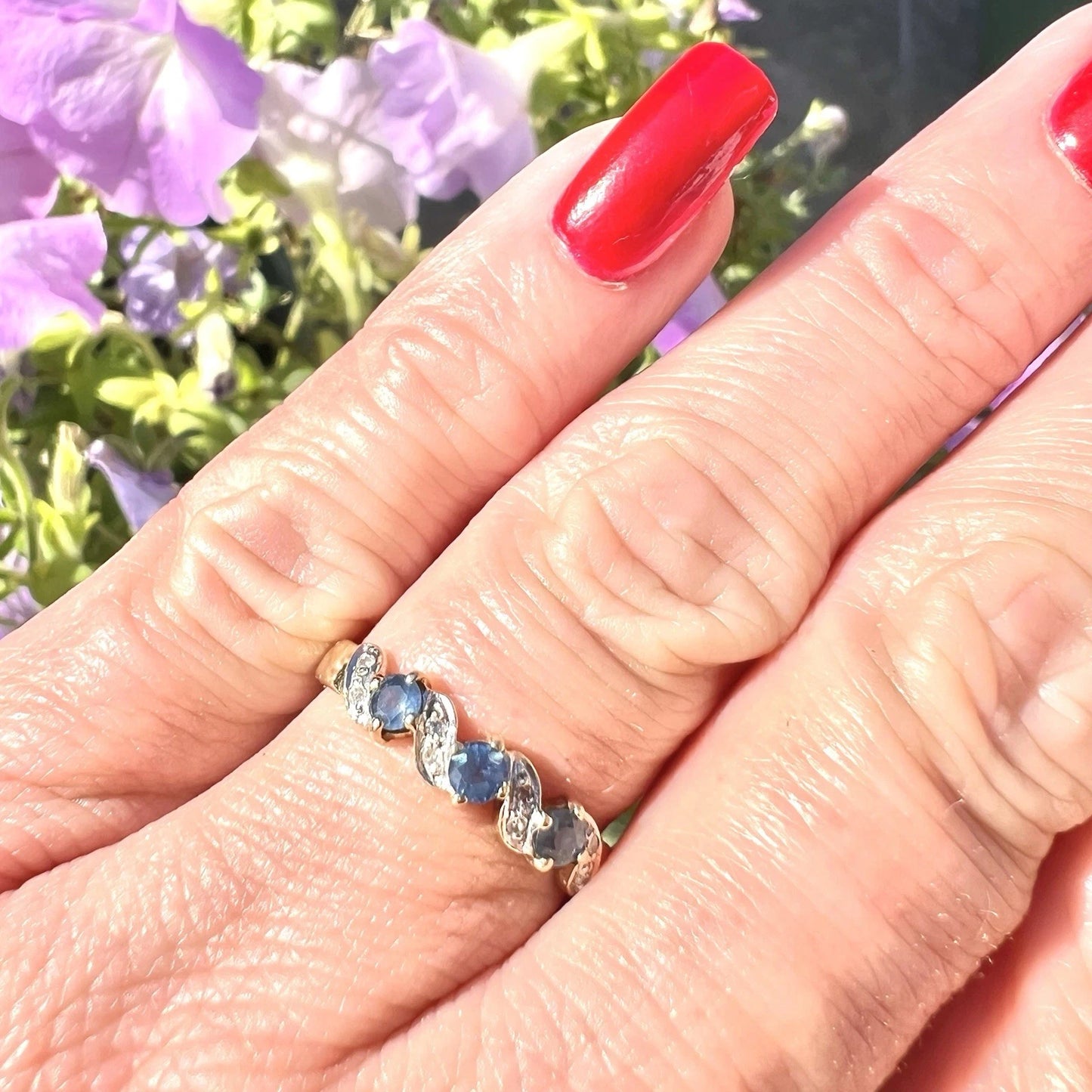 Hand wearing a ring with blue sapphire gemstones and diamonds against a blurred floral background