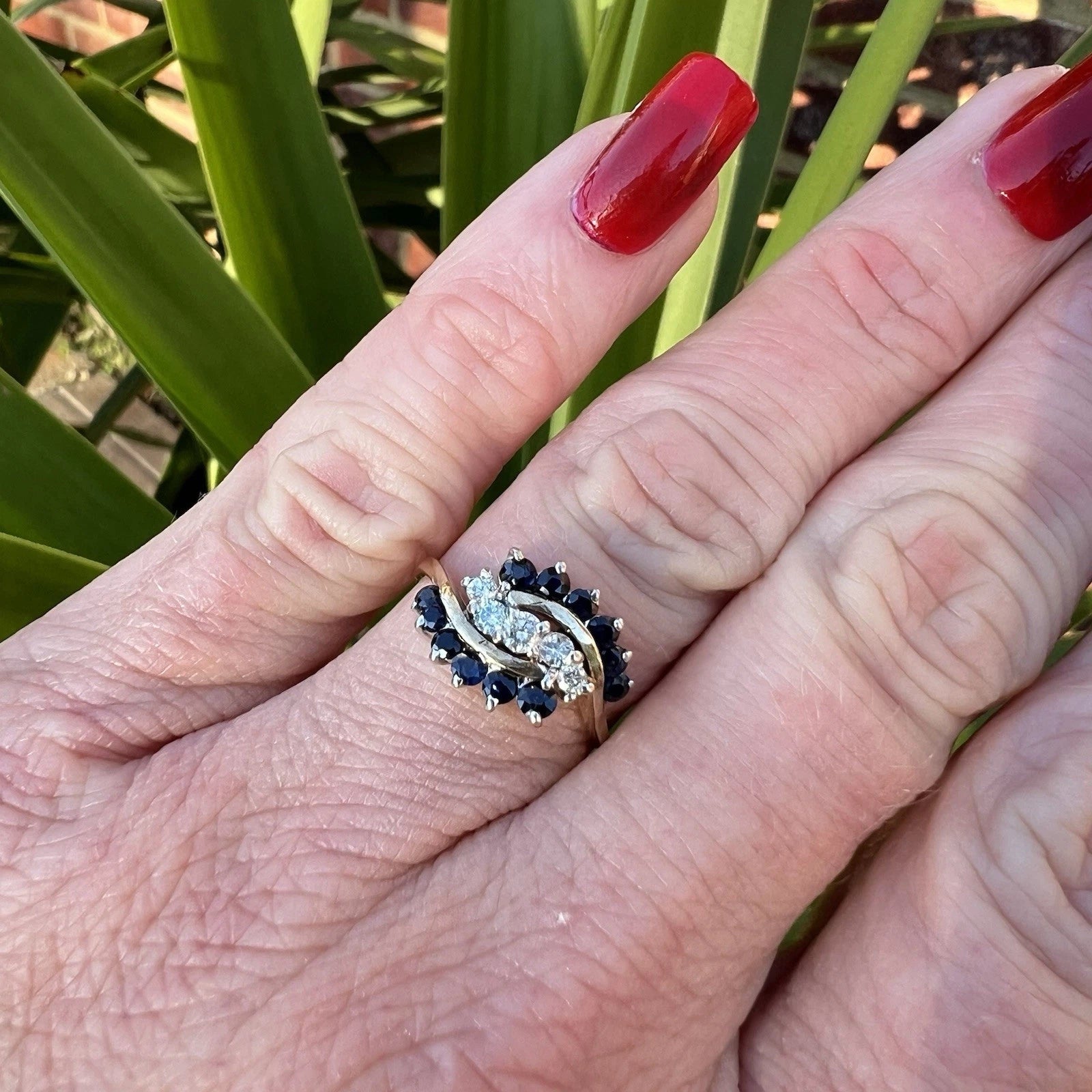 Hand wearing a ring with blue sapphire stones and diamonds against a green leafy background