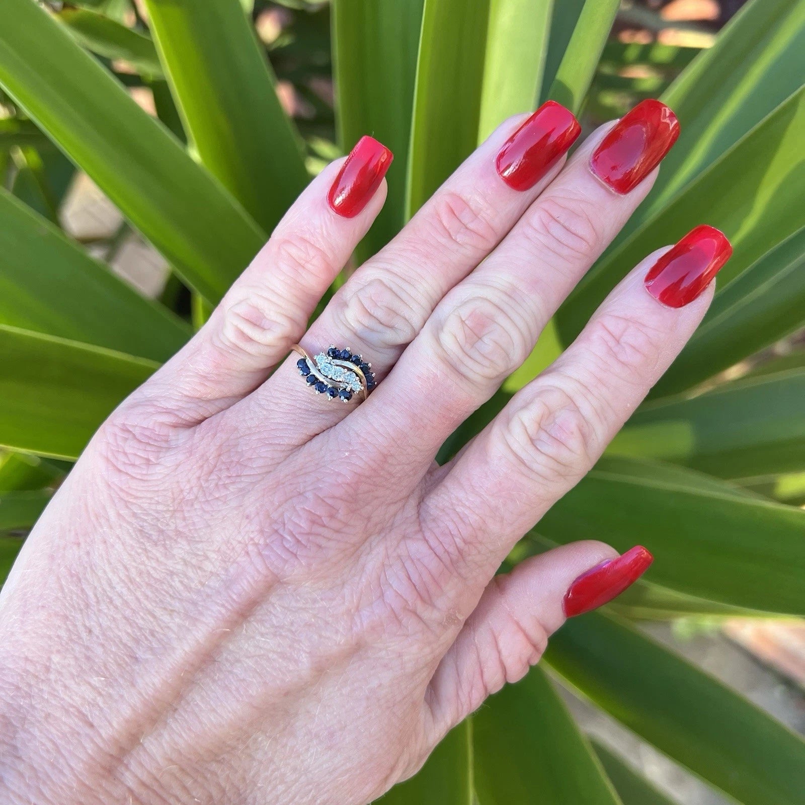 Hand with red nail polish wearing a sapphire and diamond cluster ring against a green leafy background