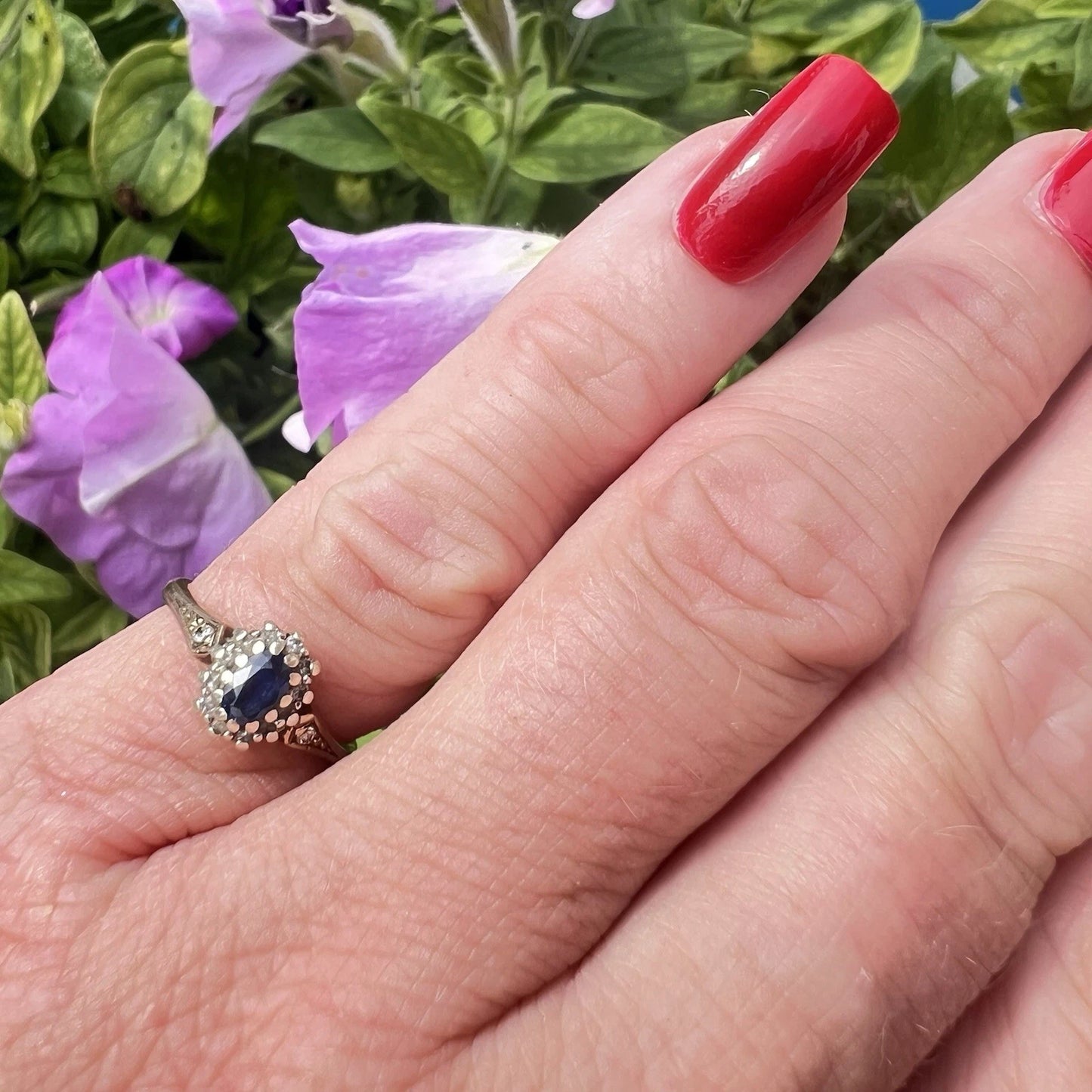 Hand with red nail polish wearing a ring with a blue sapphire gemstone against a floral background