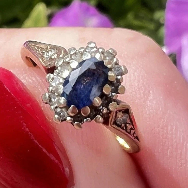 Gold ring with a blue sapphire gemstone on a finger against a blurred floral background
