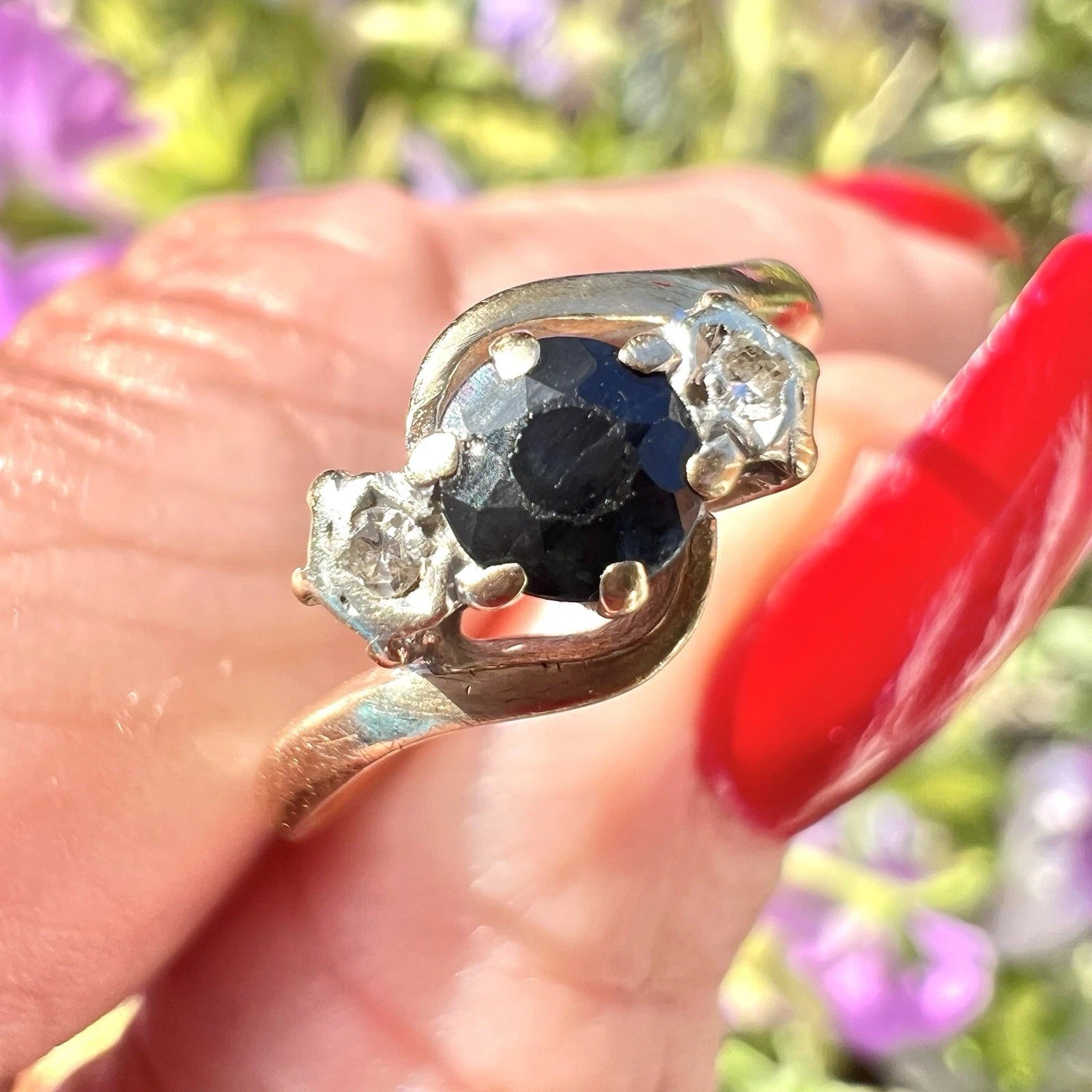 Gold ring with a sapphire gemstone and diamond held by a hand with red nail polish against a blurred natural background.