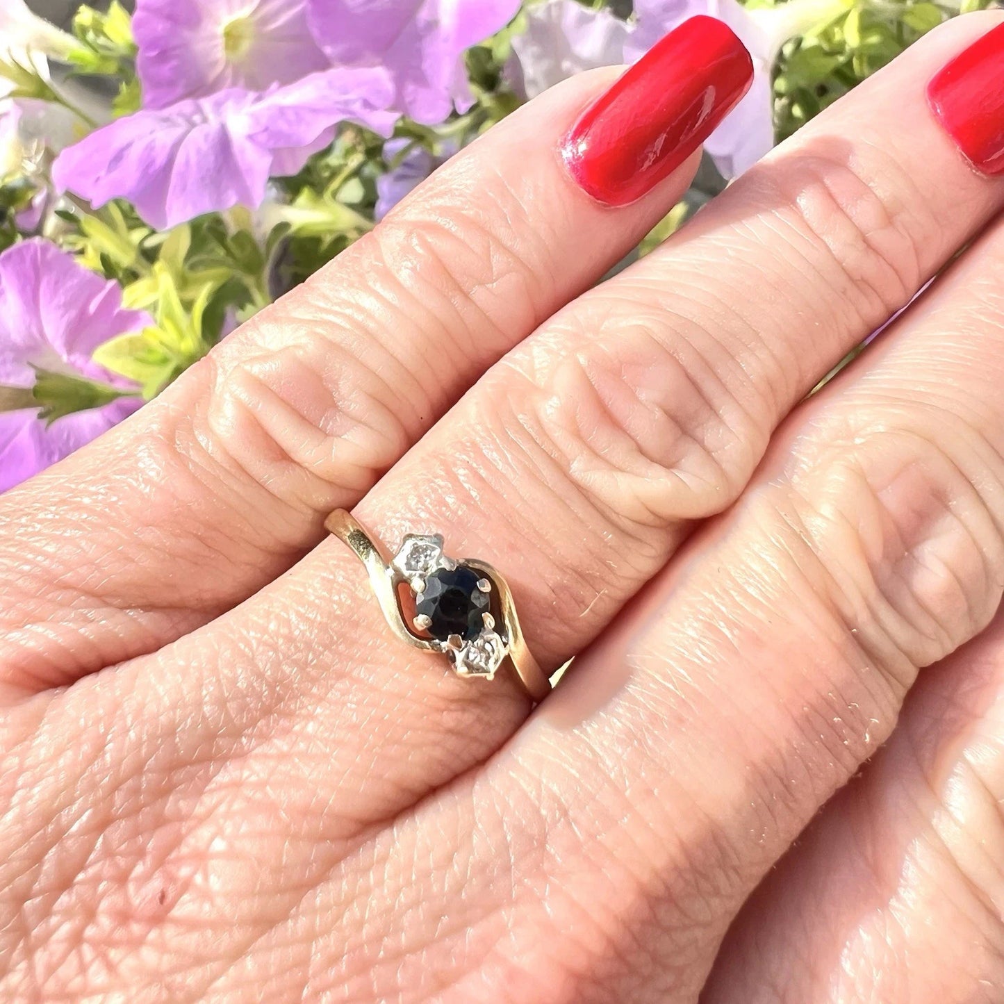 Gold ring with a sapphire and gemstone and diamonds on a hand with red nail polish against a blurred floral background.