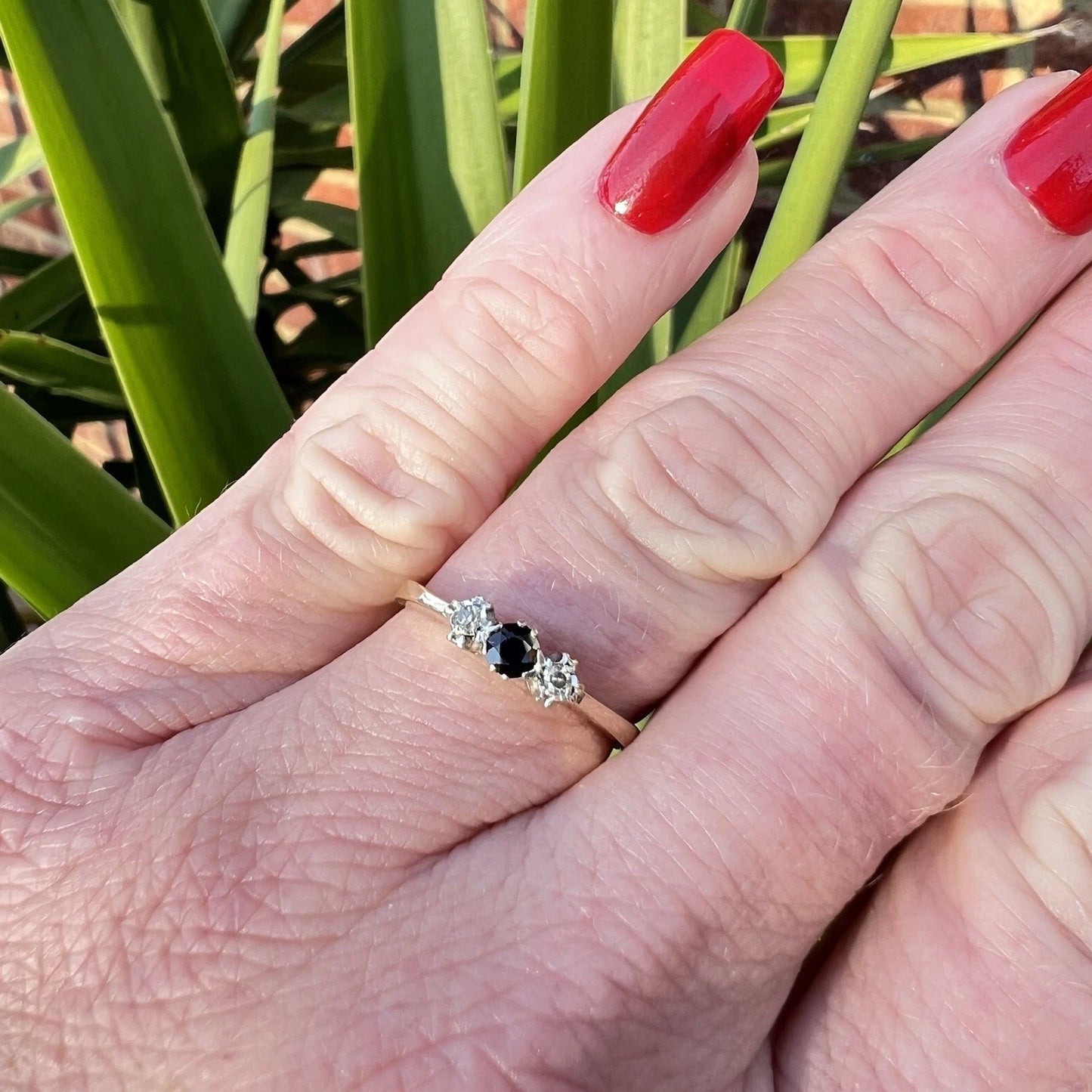 Hand wearing a ring with a sapphire and diamond accents against a green plant background