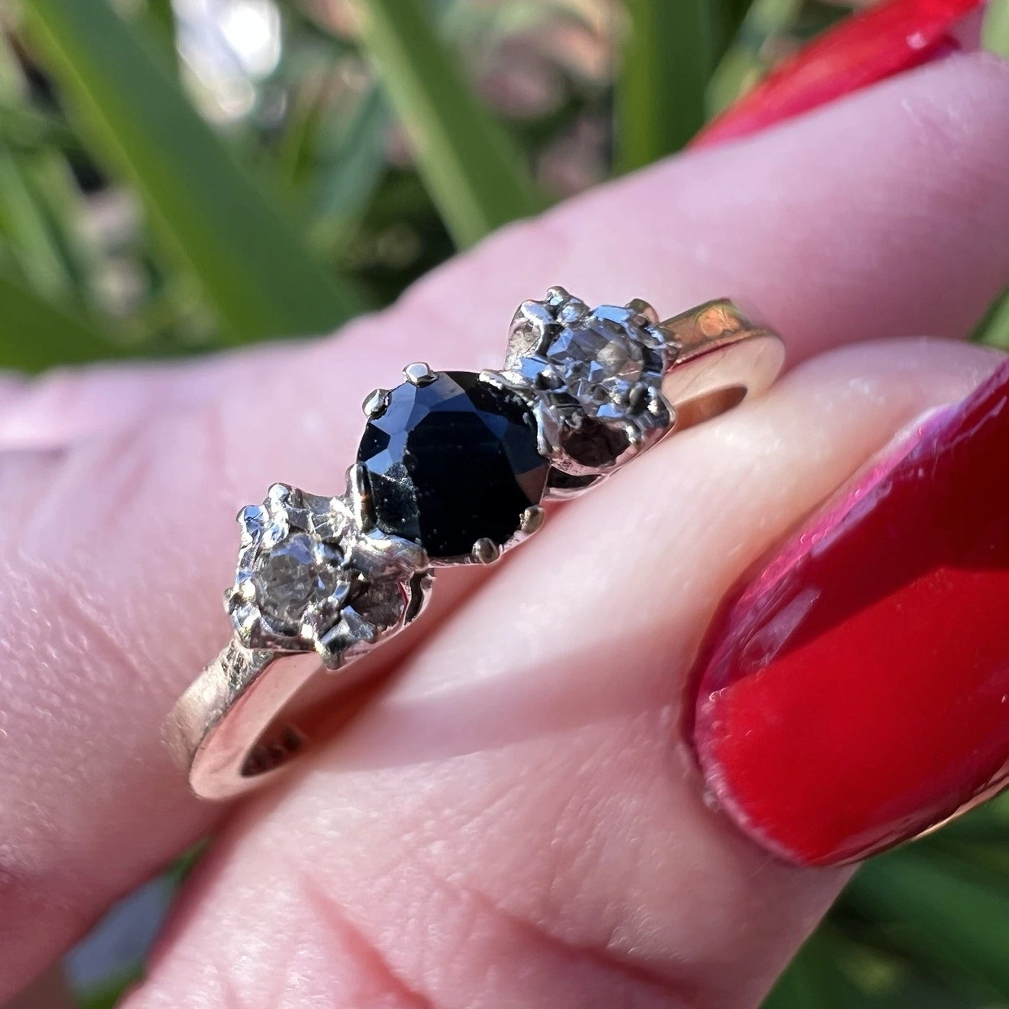 Gold ring with a sapphire and diamonds on a finger with red nail polish.