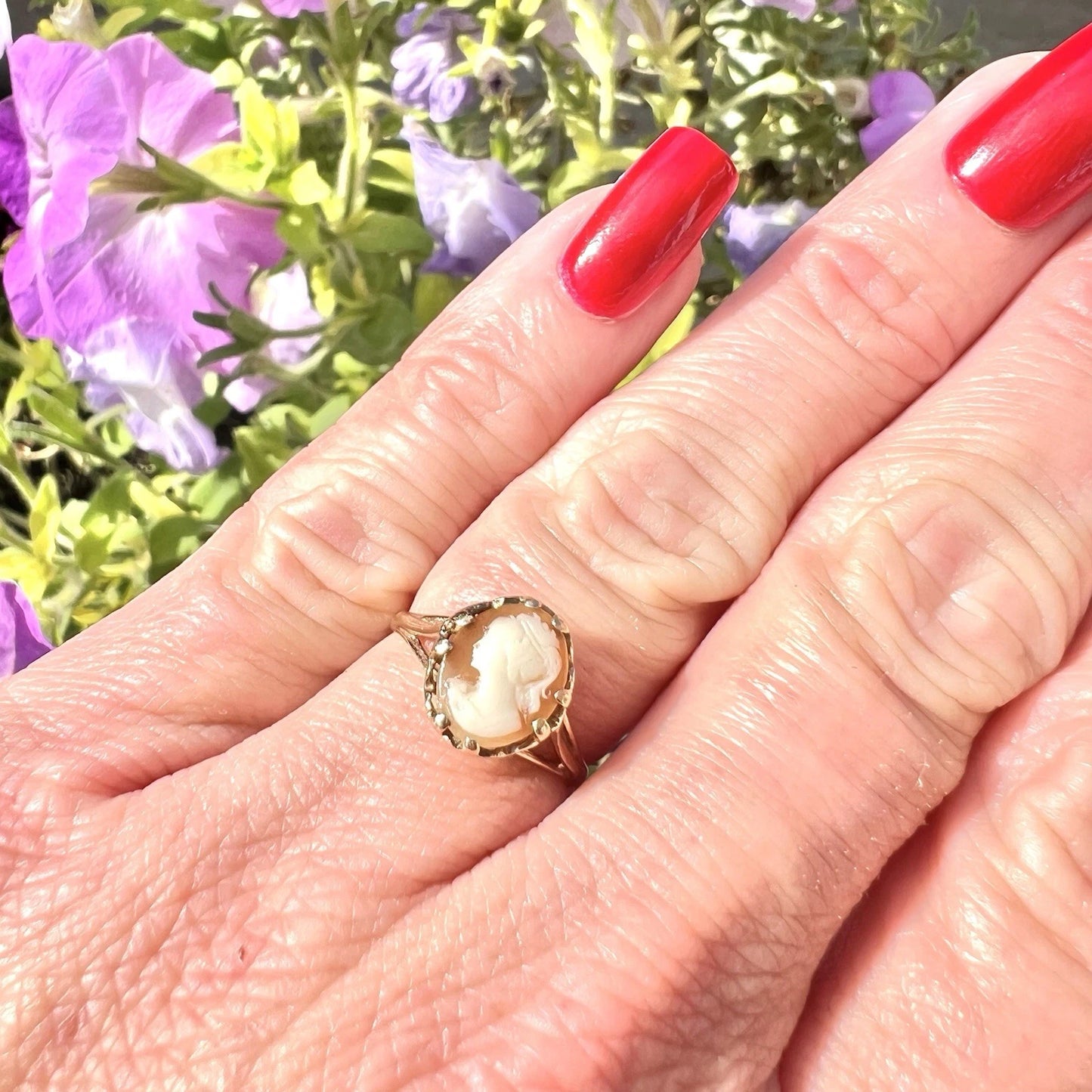Hand wearing a gold ring with a cameo design against a blurred floral background