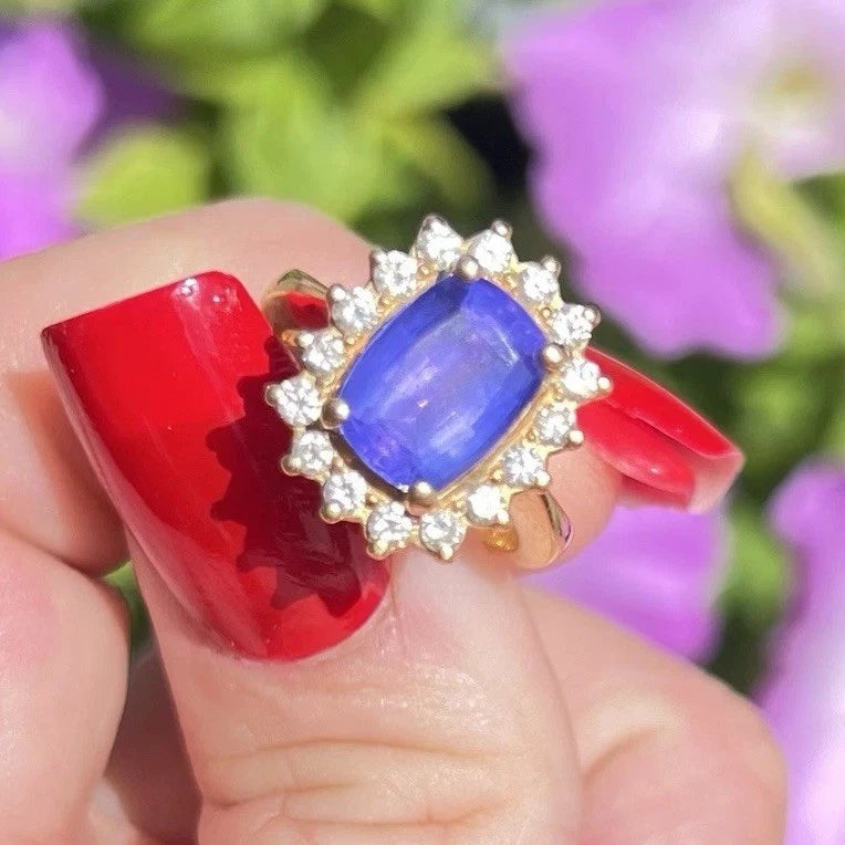 Ring with a blue tanzanite gemstone and diamond halo held by fingers with red nail polish against a blurred floral background.