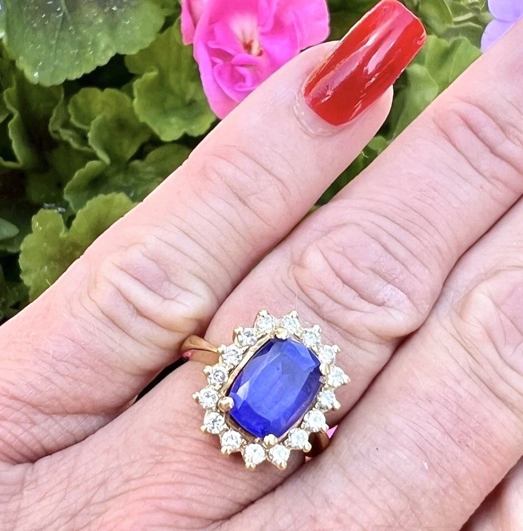 Gold ring with a blue tanzanite gemstone and diamond halo worn on a finger with red nail polish, against a floral background.