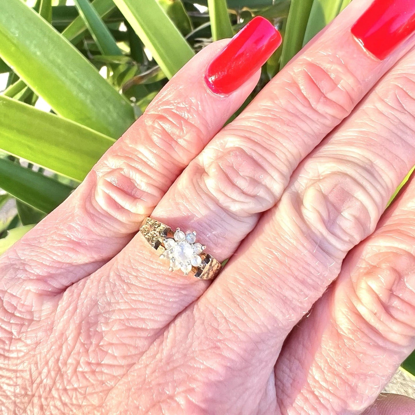 Hand wearing a gold cluster ring set with topaz, red nail polish against a green leafy background