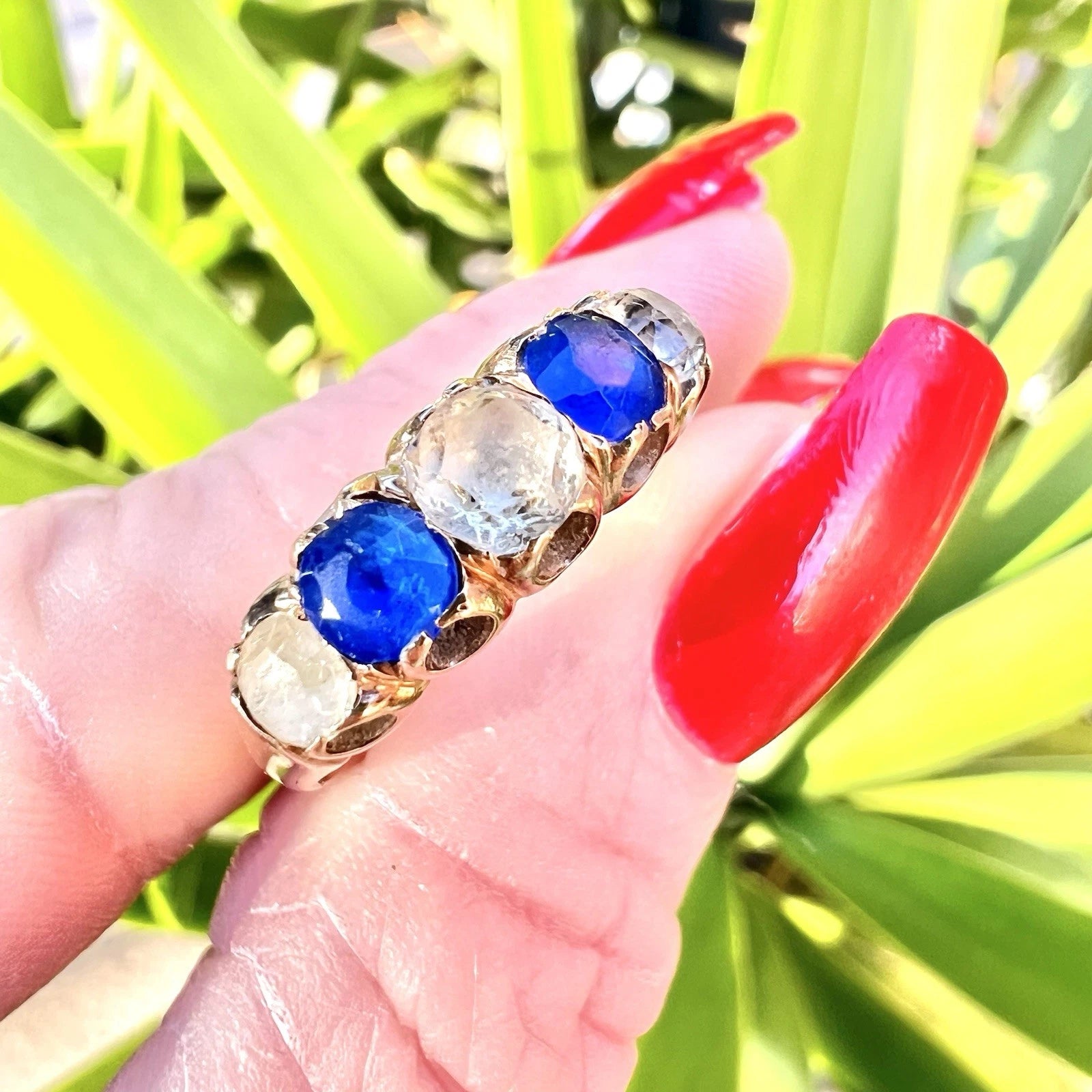 Antique ring with blue and clear gemstones on a hand with red nail polish against a green leafy background