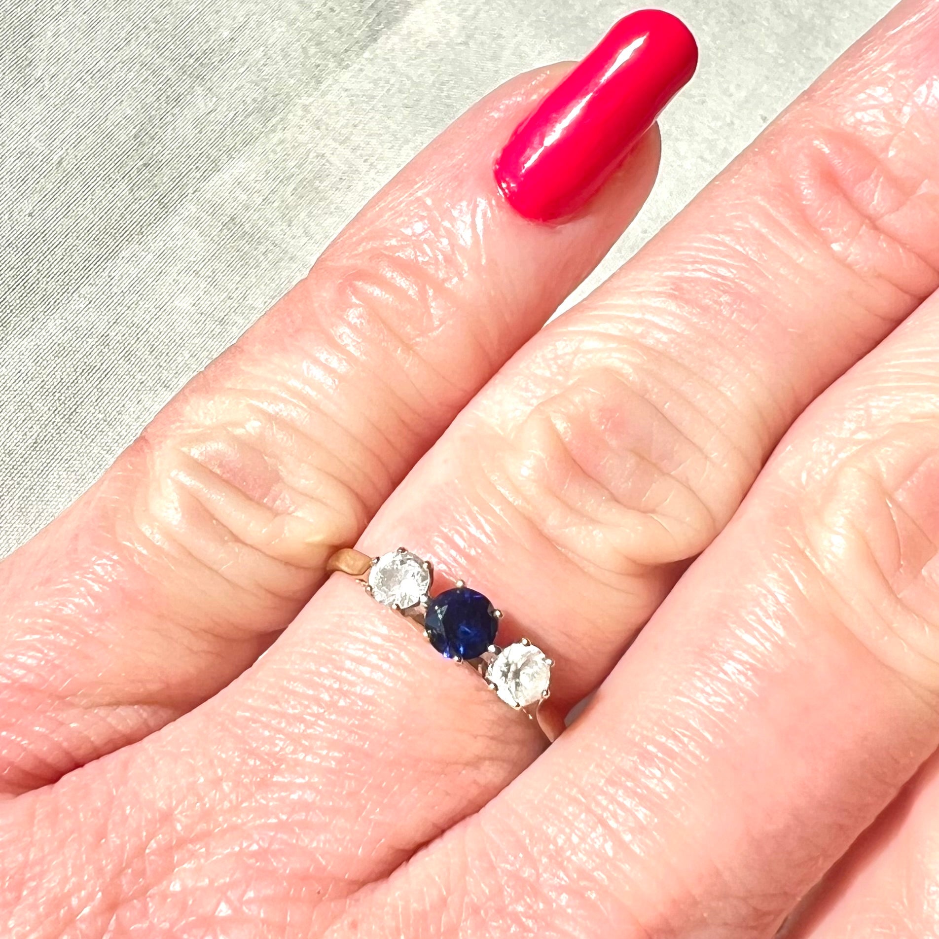 Three-stone ring with a blue gemstone and diamonds on a finger with pink nail polish.