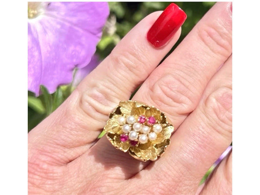 Gold ring with pearls and rubies on a hand with red nail polish, against a blurred floral background.