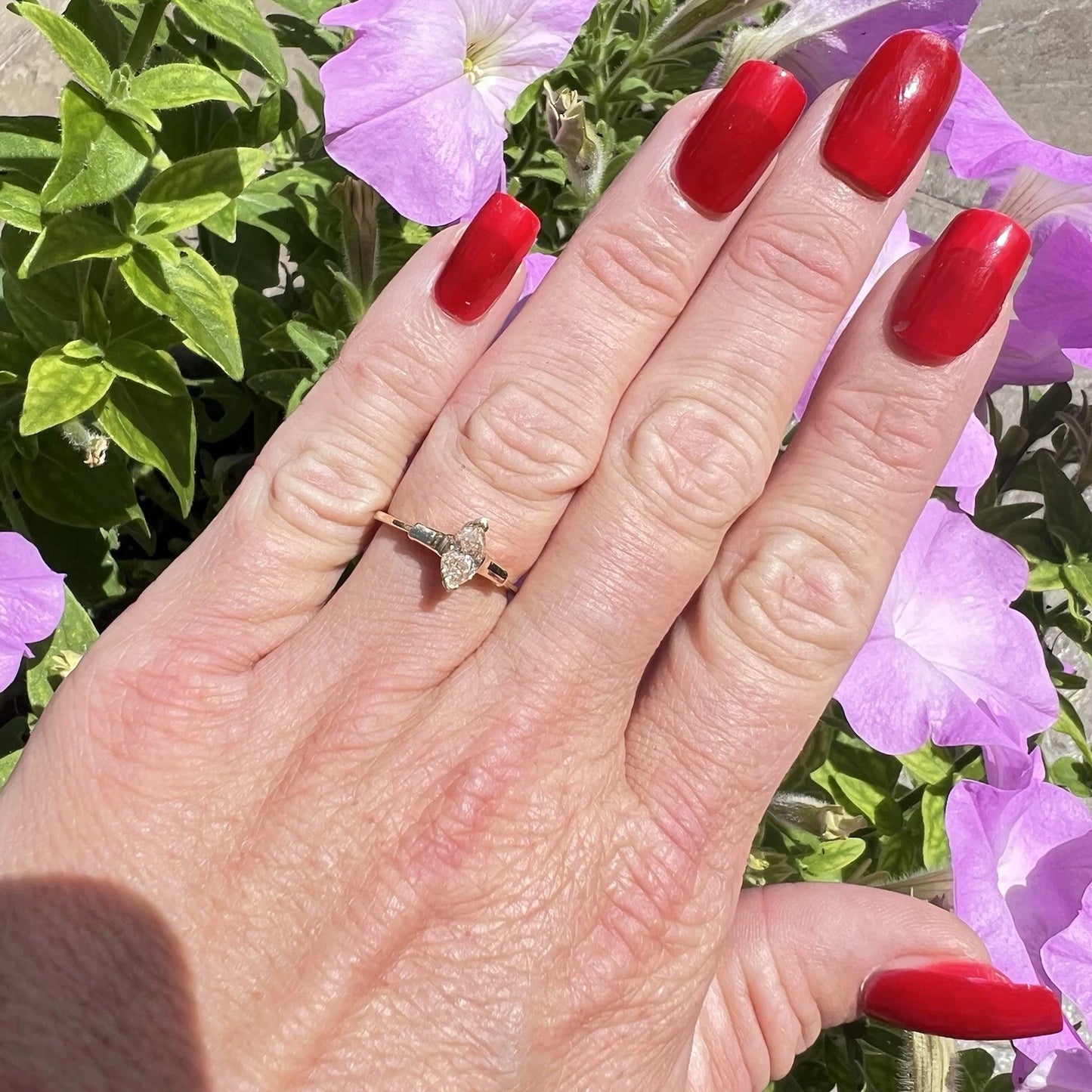 Hand with red nail polish wearing a gold yellow diamond solitaire ring against a floral background